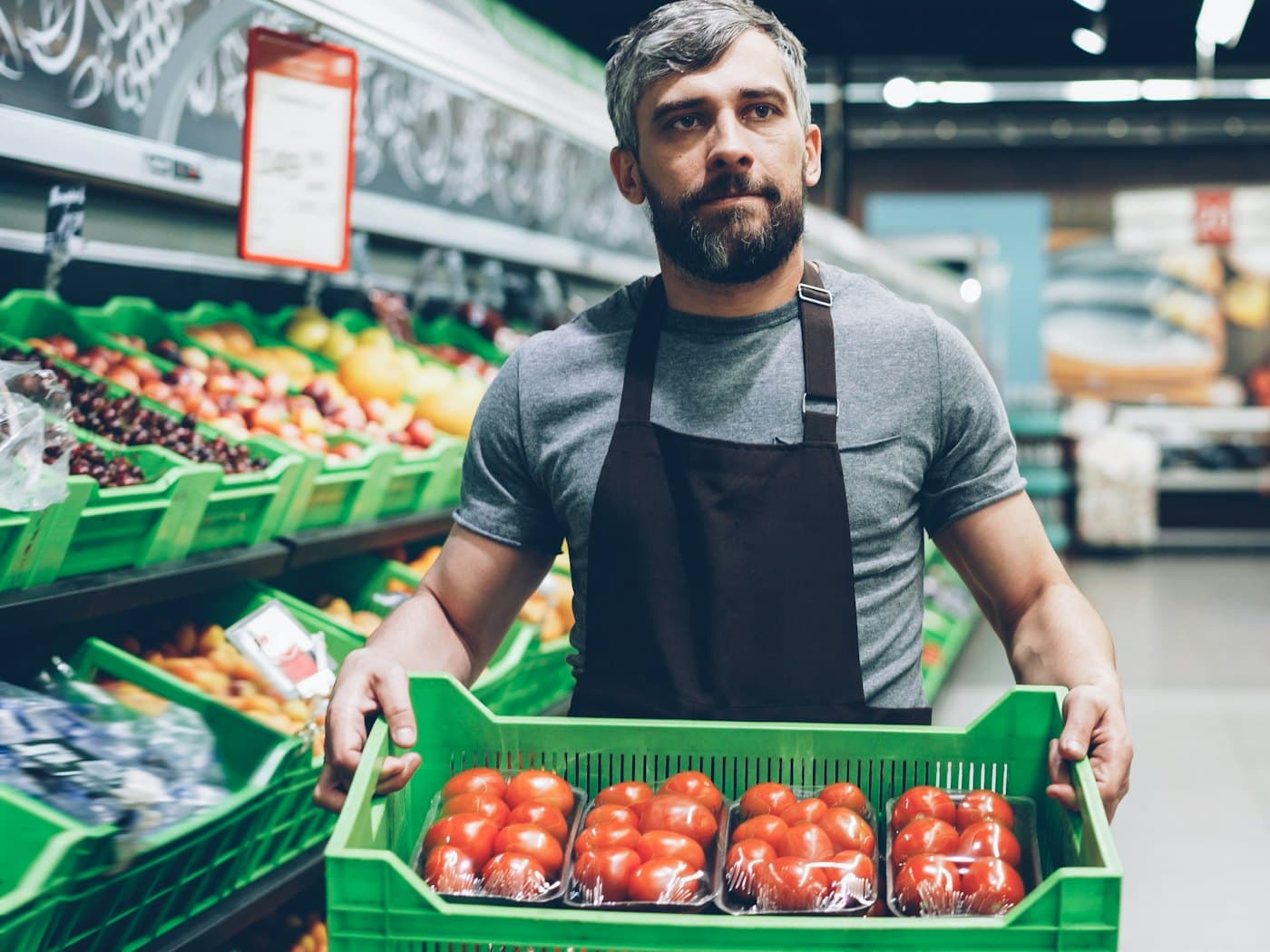 Un commerçant tient une caisse de tomates devant son rayon de fruits et légumes